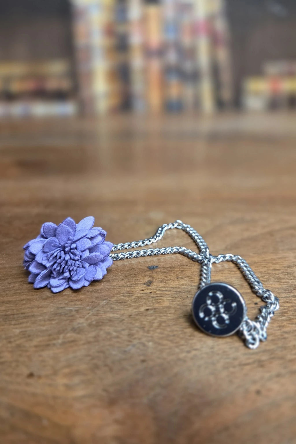 A purple flower lapel pin, silver chain and a round silver Cavani medallion pendant, resting on a wooden tabletop with a blurred background.
