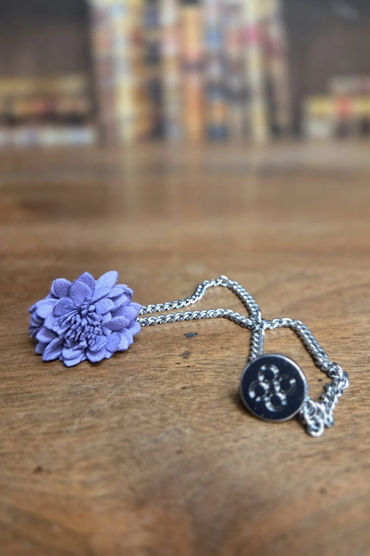 A silver chain necklace featuring a lavender-purple fabric flower on one end and a round black button charm with white embroidered details on the other, resting on a wooden surface.