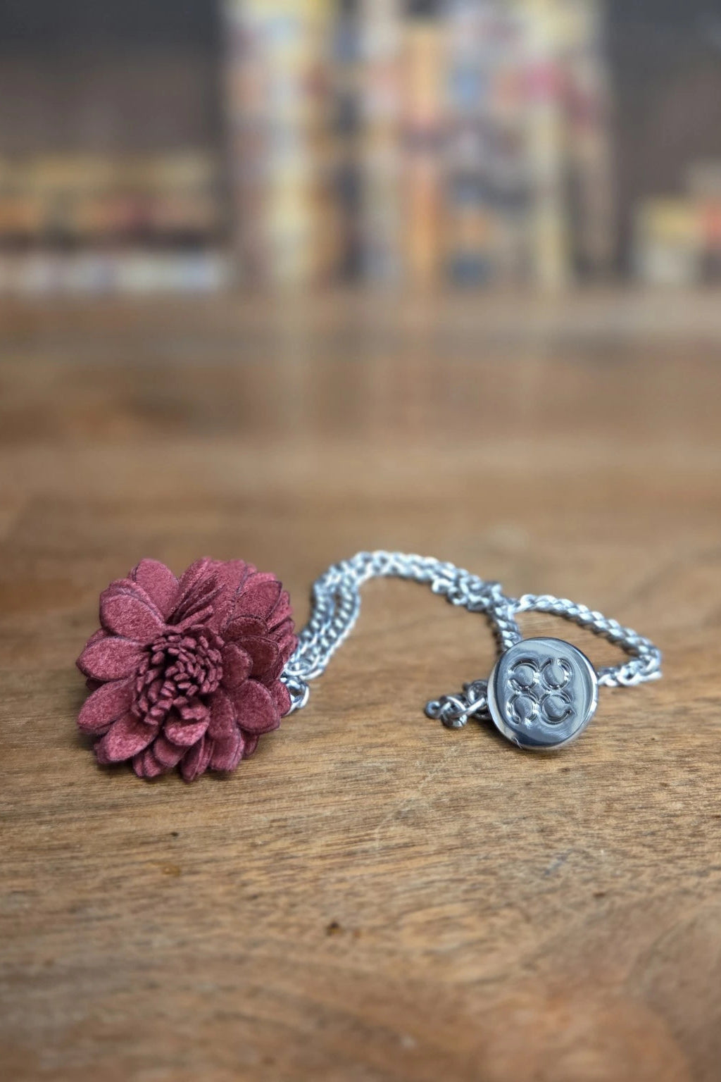 A dark burgundy flower lapel pin, silver chain and a round silver Cavani medallion pendant, resting on a wooden tabletop with a blurred background.