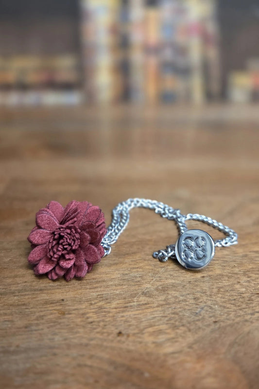 A silver chain necklace featuring a textured, dark burgundy flower charm and a round silver medallion pendant, resting on a wooden tabletop with a blurred background.