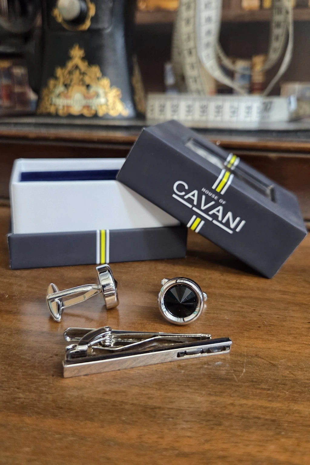 A men's formal accessory set by Cavani, including a silver-toned tie clip, a round watch with a black face, and silver cufflinks, displayed on a wooden table next to their branded dark gray gift box.