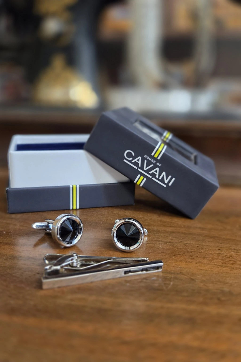 A men's formal accessory set including round silver cufflinks with black faces, a silver tie clip, and branded CAVANI gift boxes, arranged on a wooden table.
