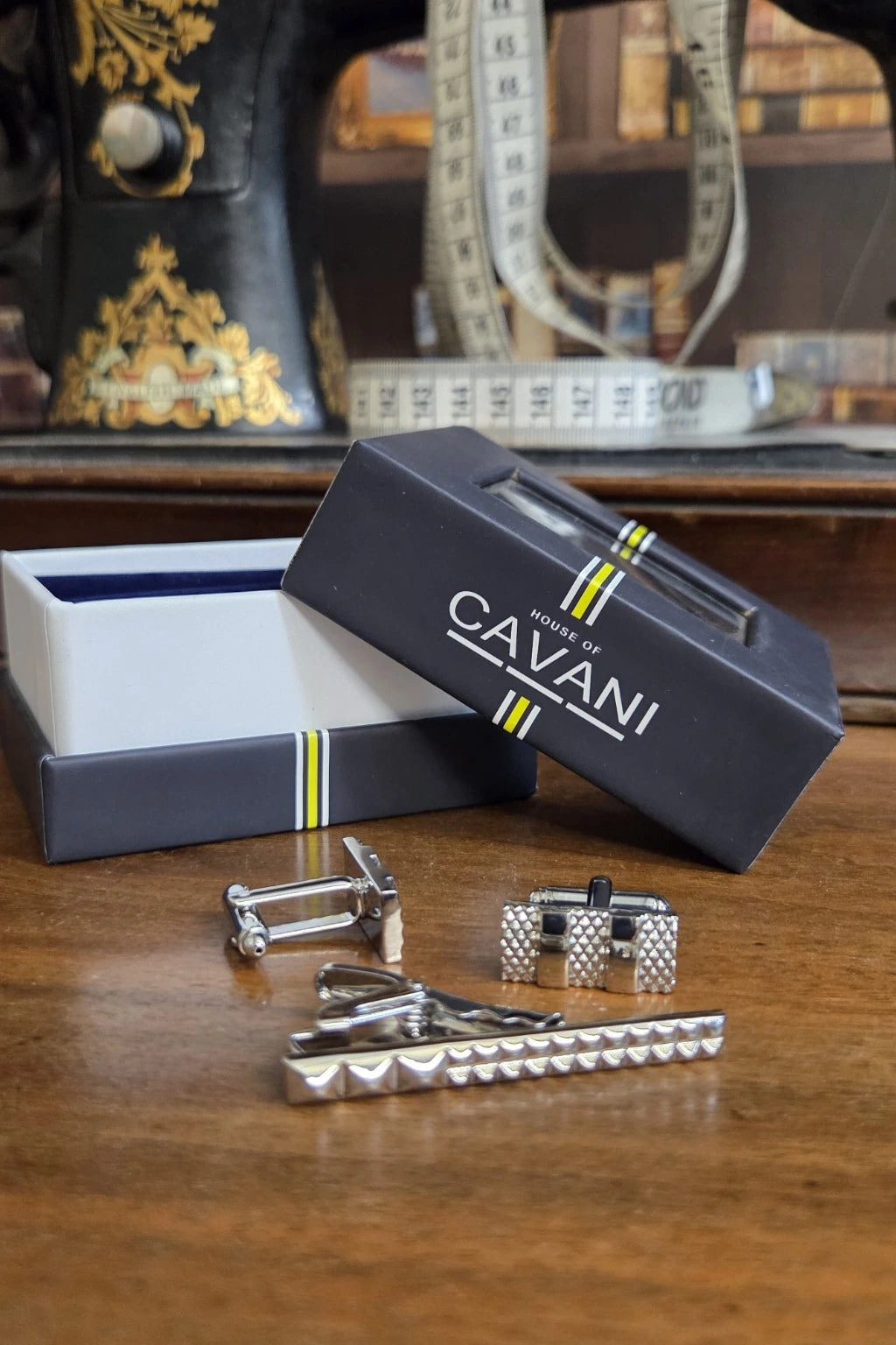 A set of silver-toned men's formal accessories, including cufflinks, a tie clip, and a matching tie bar, displayed in front of their dark navy blue CAVANI brand packaging on a wooden table.