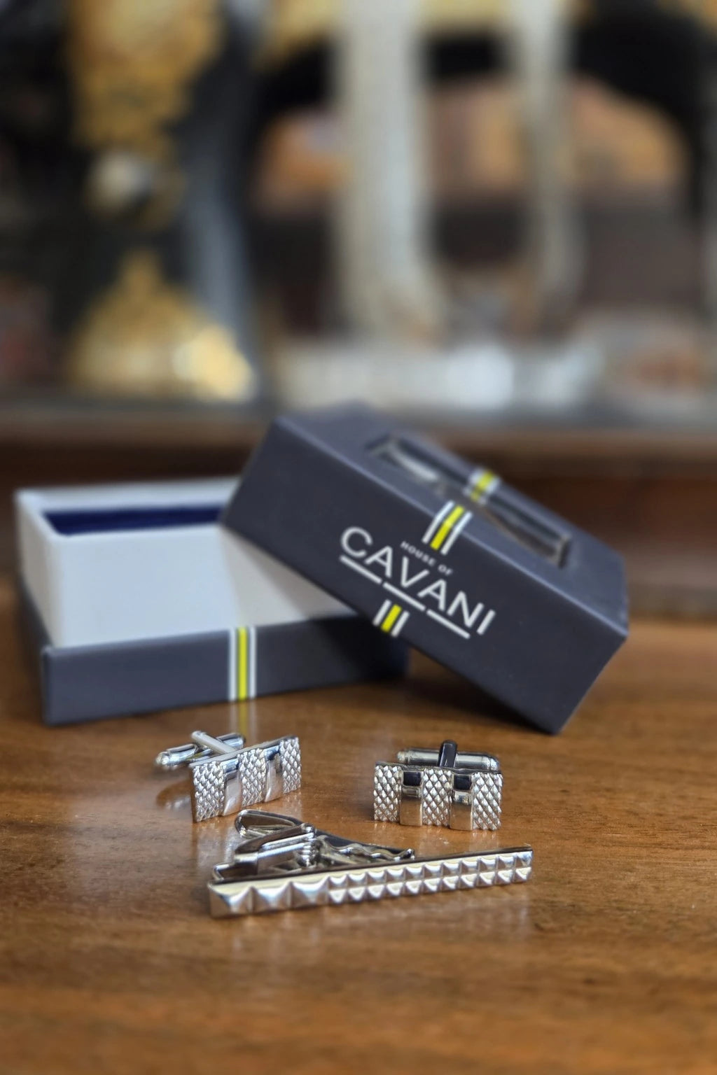 A men's formal accessory set including textured silver cufflinks, a matching silver tie clip, and a dark navy Cavani gift box on a wooden table.