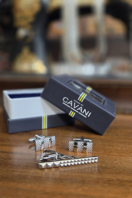A men's formal accessory set including textured silver cufflinks, a matching silver tie clip, and a dark navy Cavani gift box on a wooden table.