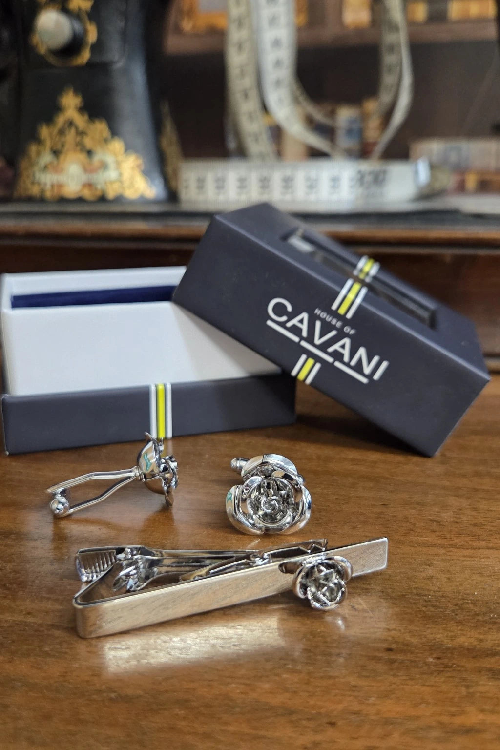 A silver-toned tie clip and matching tie clipper set with flower-shaped ties, displayed on a wooden table next to an open box and a branded black CAVANI gift box.