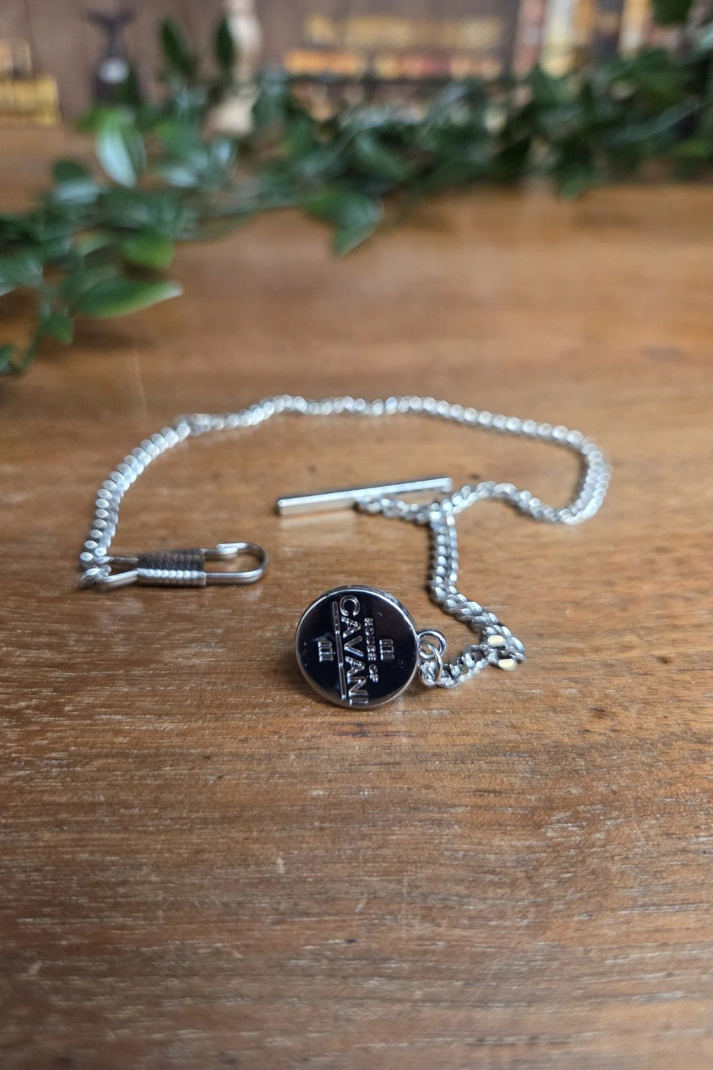 A silver-toned Cavani t-bar chain with a toggle clasp and a round silver toned pendant engraved with 'Cavani logo, displayed on a wooden tabletop with blurred greenery in the background.