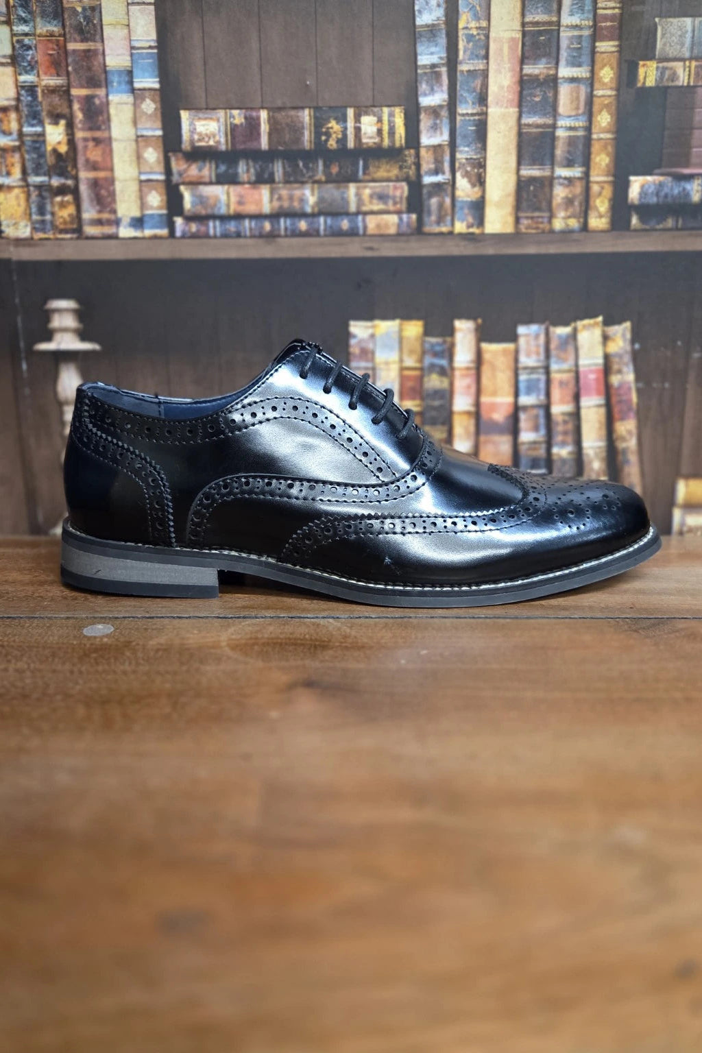 A single black leather wingtip brogue shoe with decorative perforations and a low stacked heel, displayed on a wooden surface with a blurred background of bookshelves.