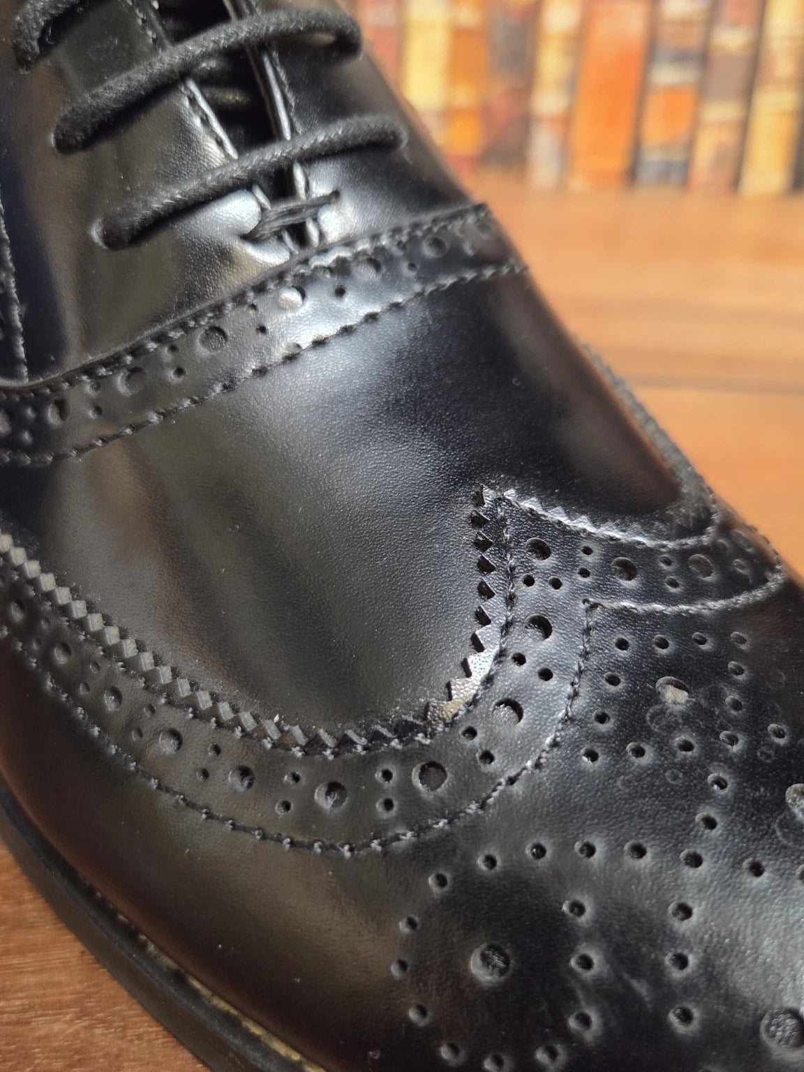 A close-up shot of a black leather wingtip oxford shoe with decorative brogue perforations and black laces, resting on a wooden surface with a blurred background.