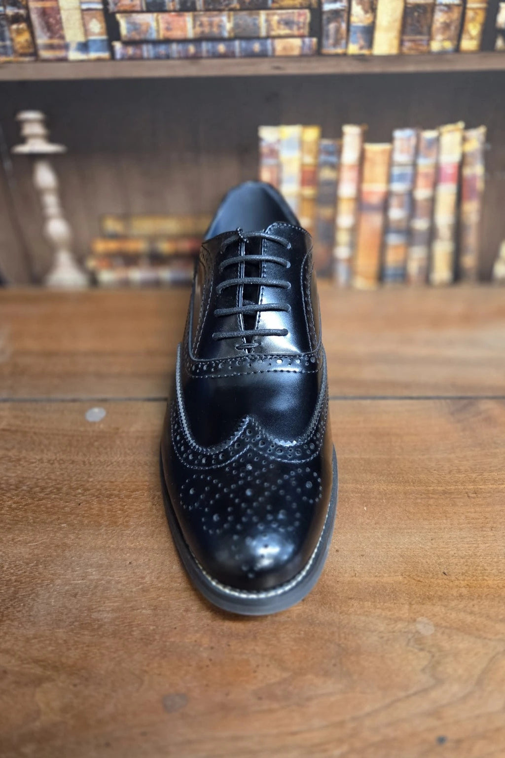 A single black leather wingtip brogue shoe with decorative perforations and laces, photographed from the front on a wooden tabletop with a blurred bookshelf background.