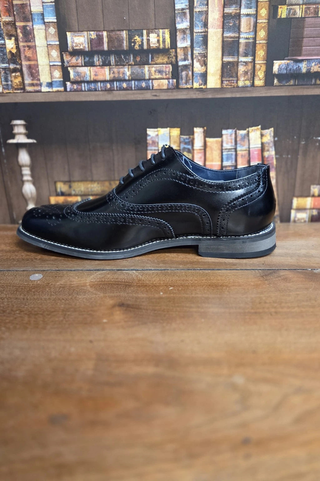 A single black leather wingtip brogue shoe with decorative stitching and a dark sole, displayed on a wooden table with a bookshelf background.