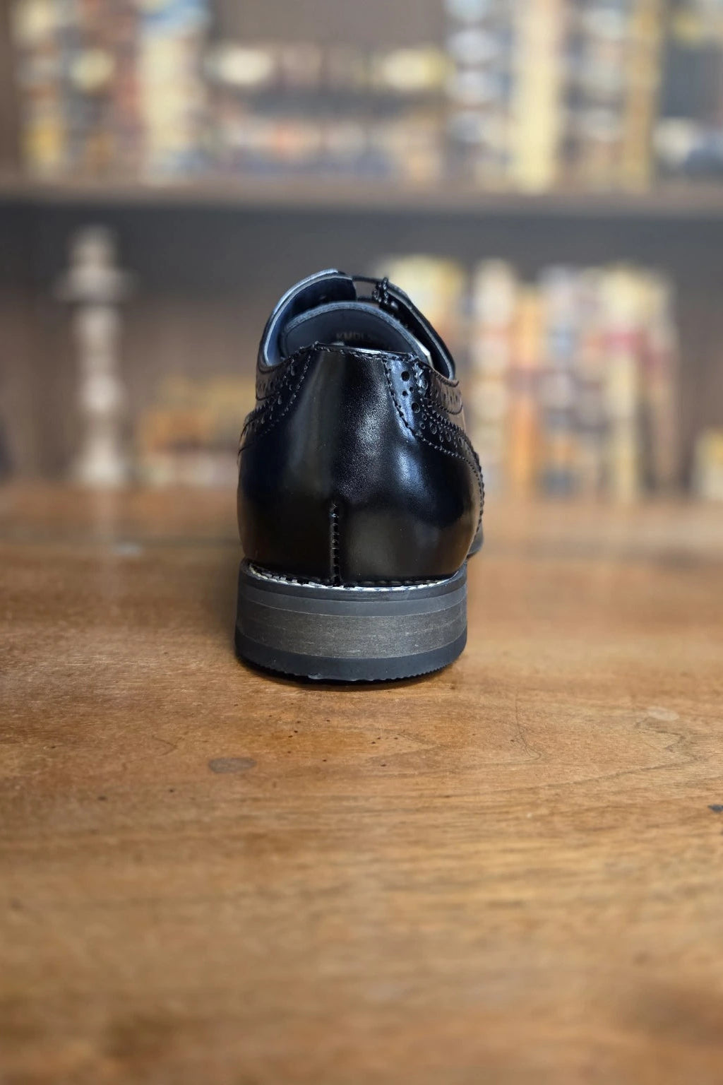 A single black leather lace-up dress shoe shown from the back, resting on a wooden tabletop with a blurred bookshelf background.