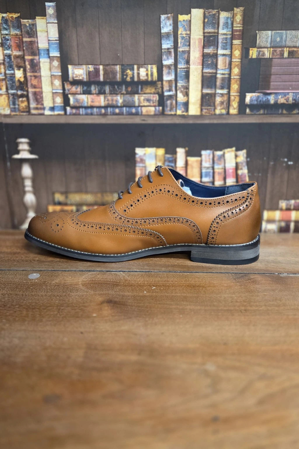 A single tan leather wingtip oxford shoe with brogue detailing, displayed in profile on a wooden table with a blurred bookshelf background.