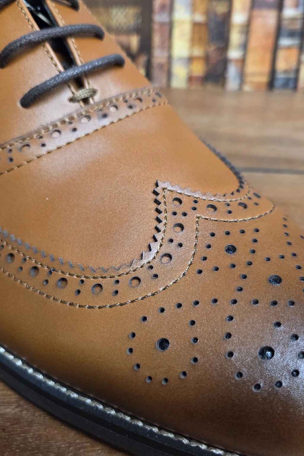 A close-up shot of a tan leather wingtip oxford shoe with decorative brogue perforations and dark laces, resting on a wooden surface with a blurred background.