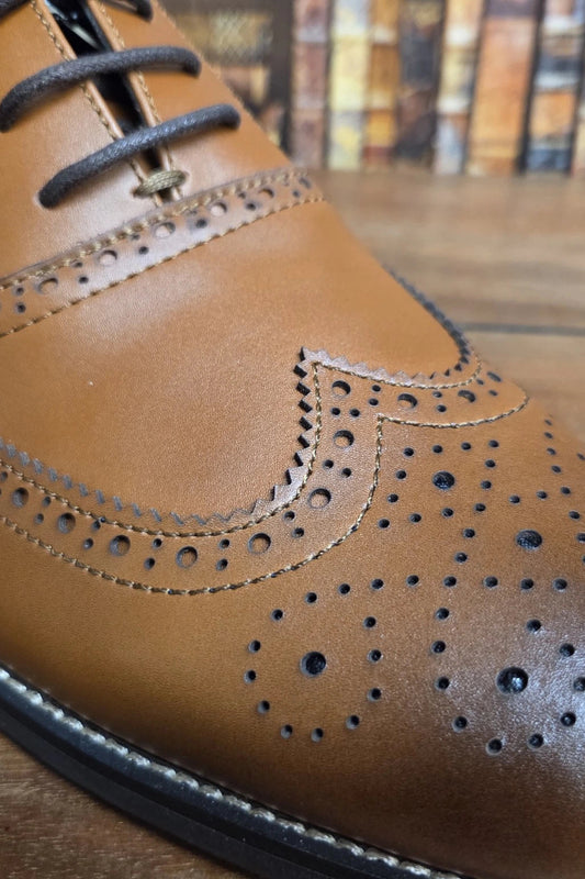 A close-up shot of a tan leather wingtip oxford shoe with decorative brogue perforations and dark laces, resting on a wooden surface with a blurred background.