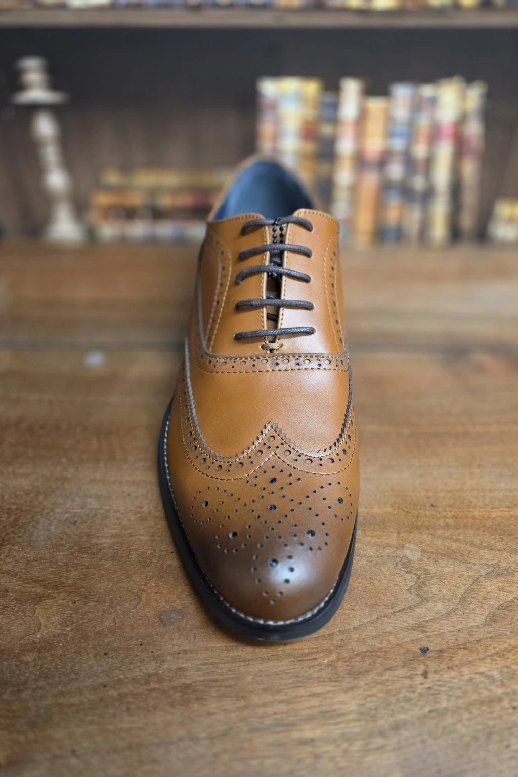 A single men's wingtip oxford shoe in tan brown leather with dark laces, photographed standing upright on a wooden tabletop with a blurred bookshelf background.