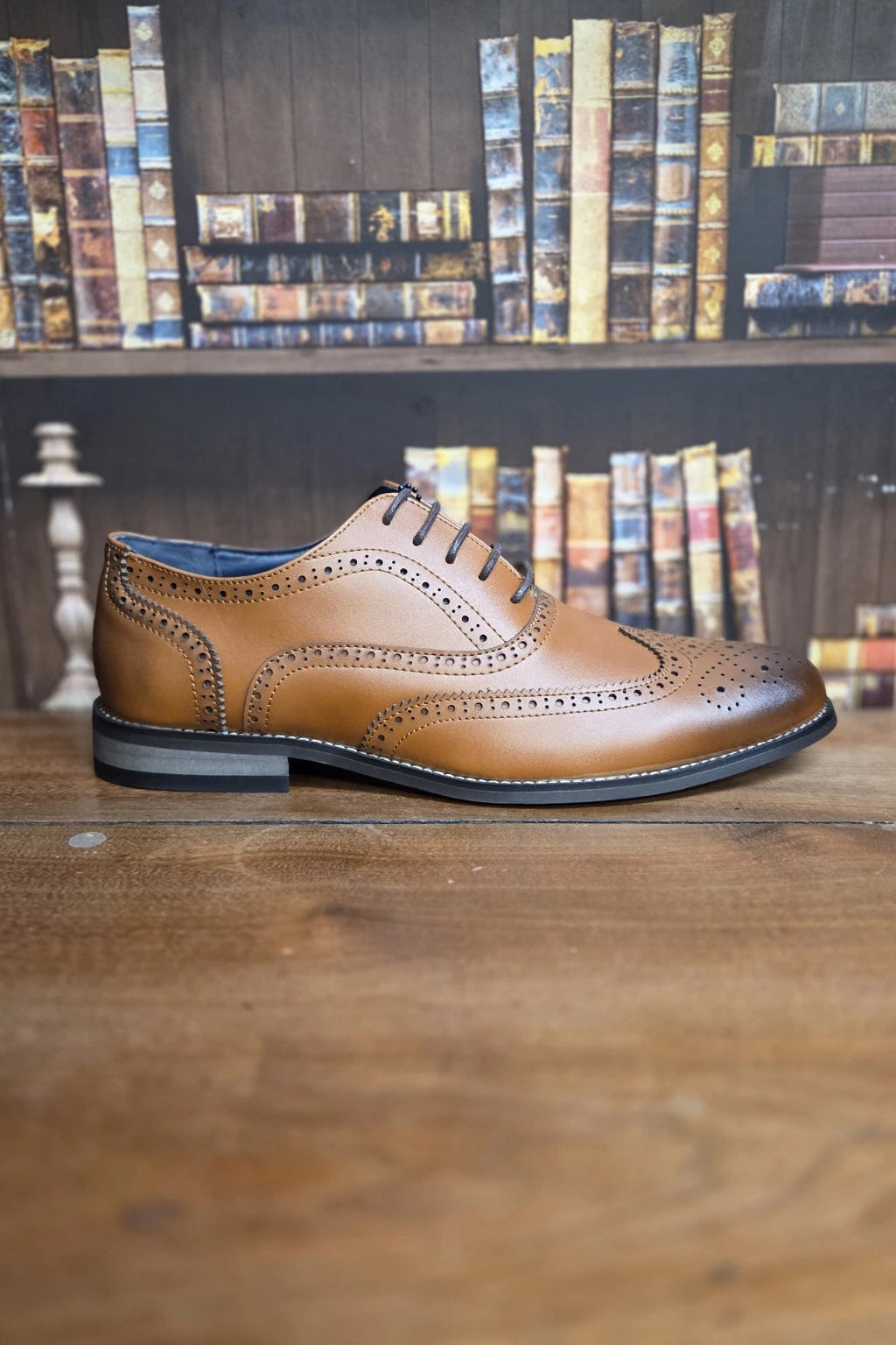 A single tan leather wingtip oxford shoe with decorative perforations and brogue detailing, displayed on a wooden table with a blurred bookshelf background.