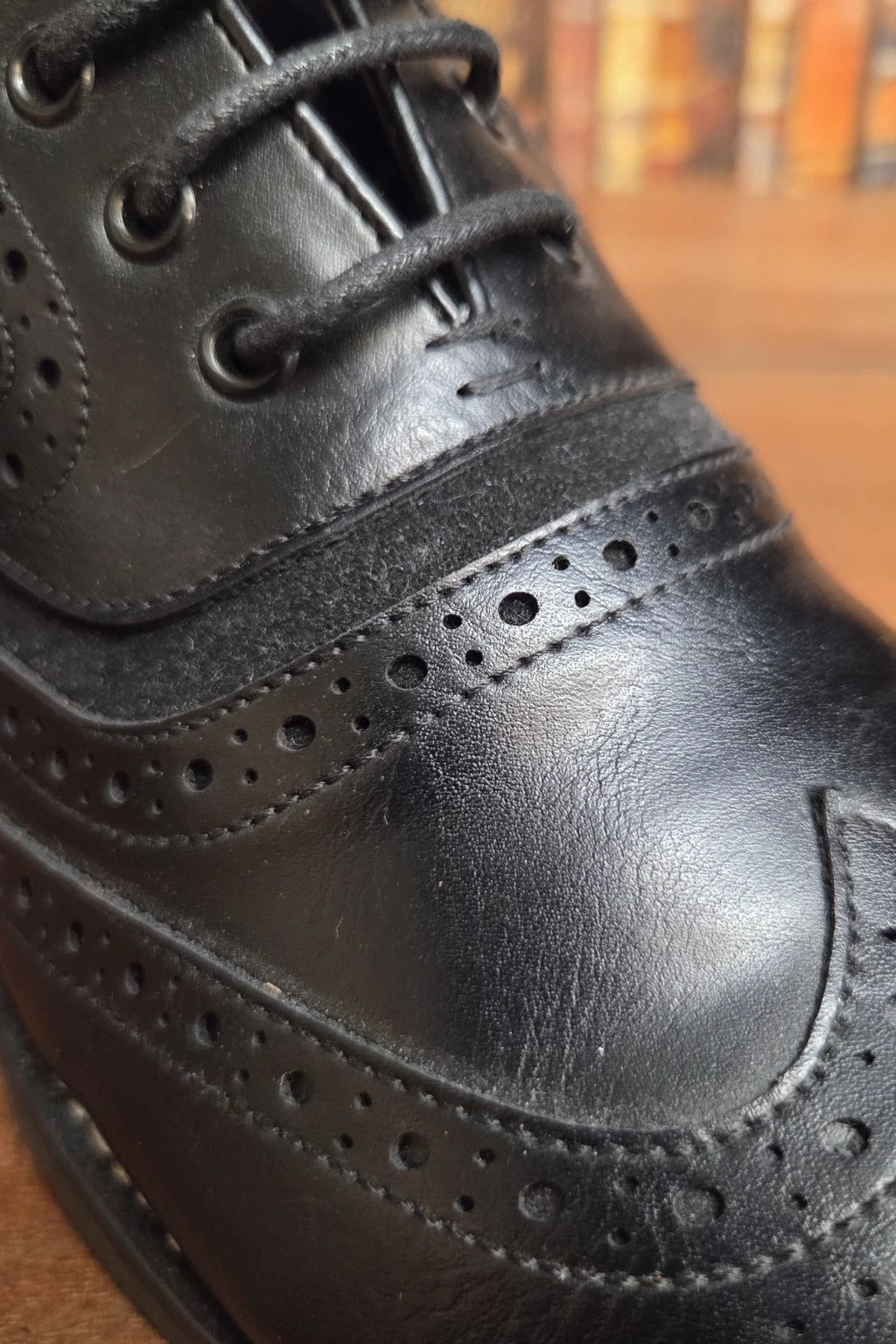 A close-up shot of a black leather wingtip brogue dress shoe, showing the detailed brogue perforations, laces, and leather texture against a warm wooden background.