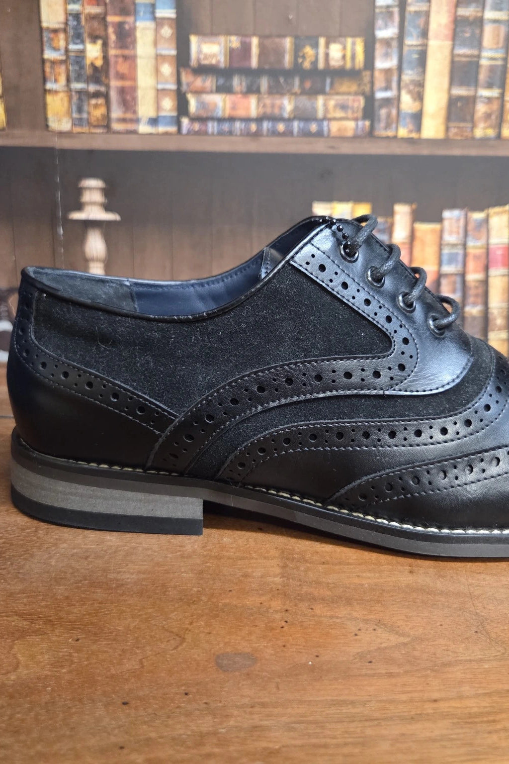 A black leather and suede wingtip brogue shoe with a low stacked heel, resting on a wooden table with a bookshelf in the background.