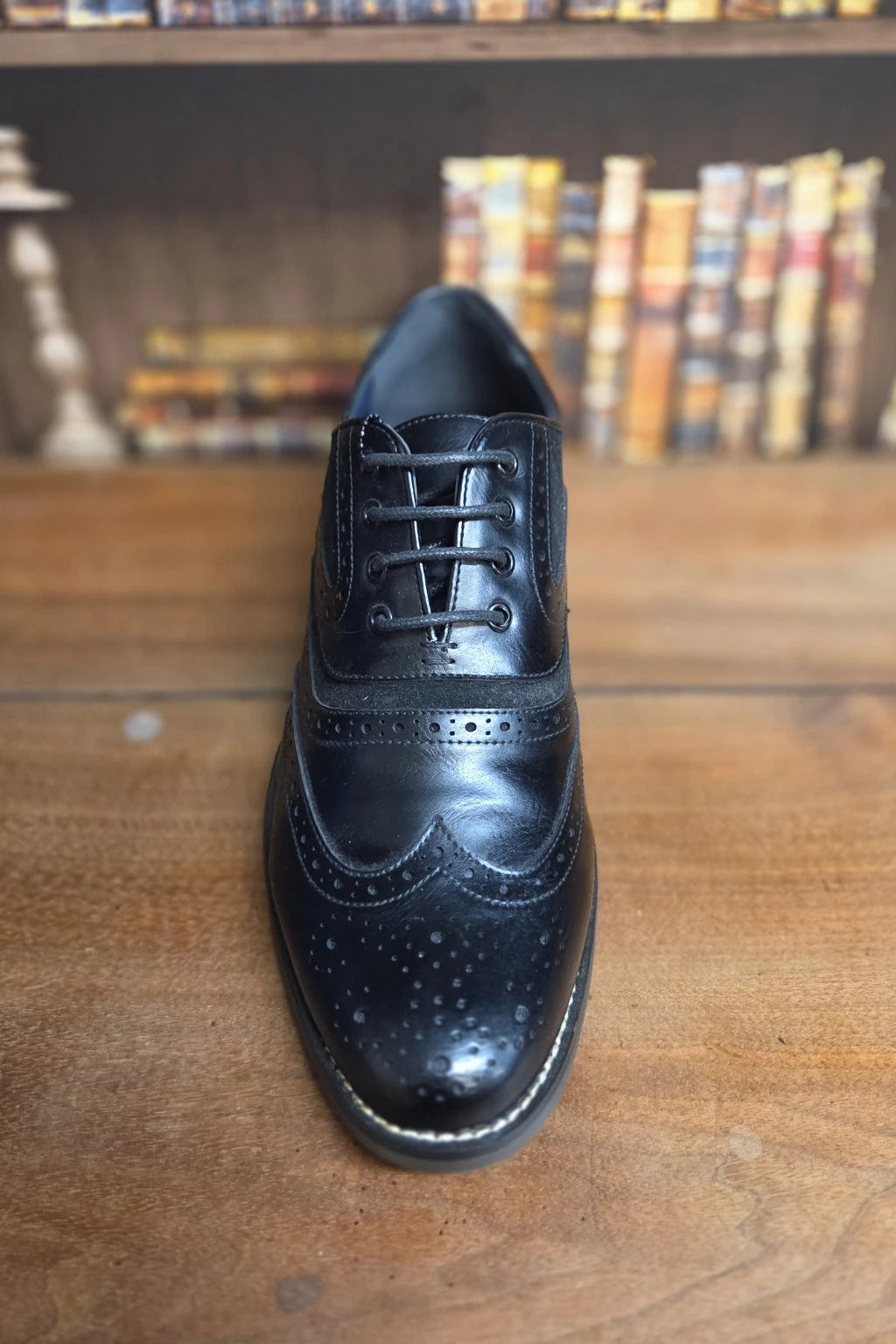 A single black leather wingtip oxford shoe with decorative brogue detailing and white contrast stitching, positioned on a wooden tabletop with a blurred background of bookshelves.
