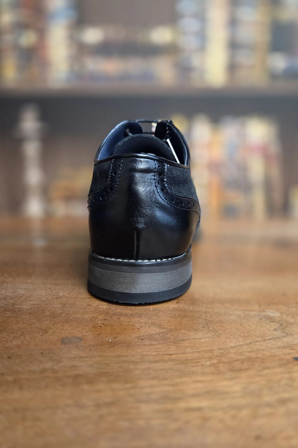 A black leather dress shoe shown from the back, resting on a wooden table with a blurred bookshelf background.