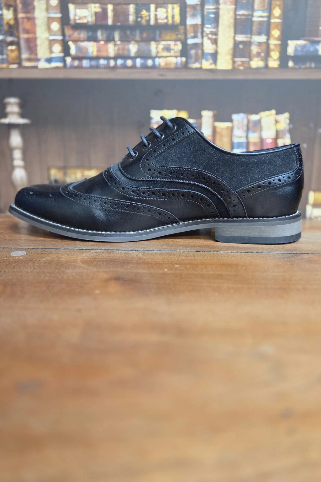 A single black leather wingtip brogue shoe with decorative brogue detailing and a low stacked heel, positioned on a wooden tabletop with a blurred bookshelf background.