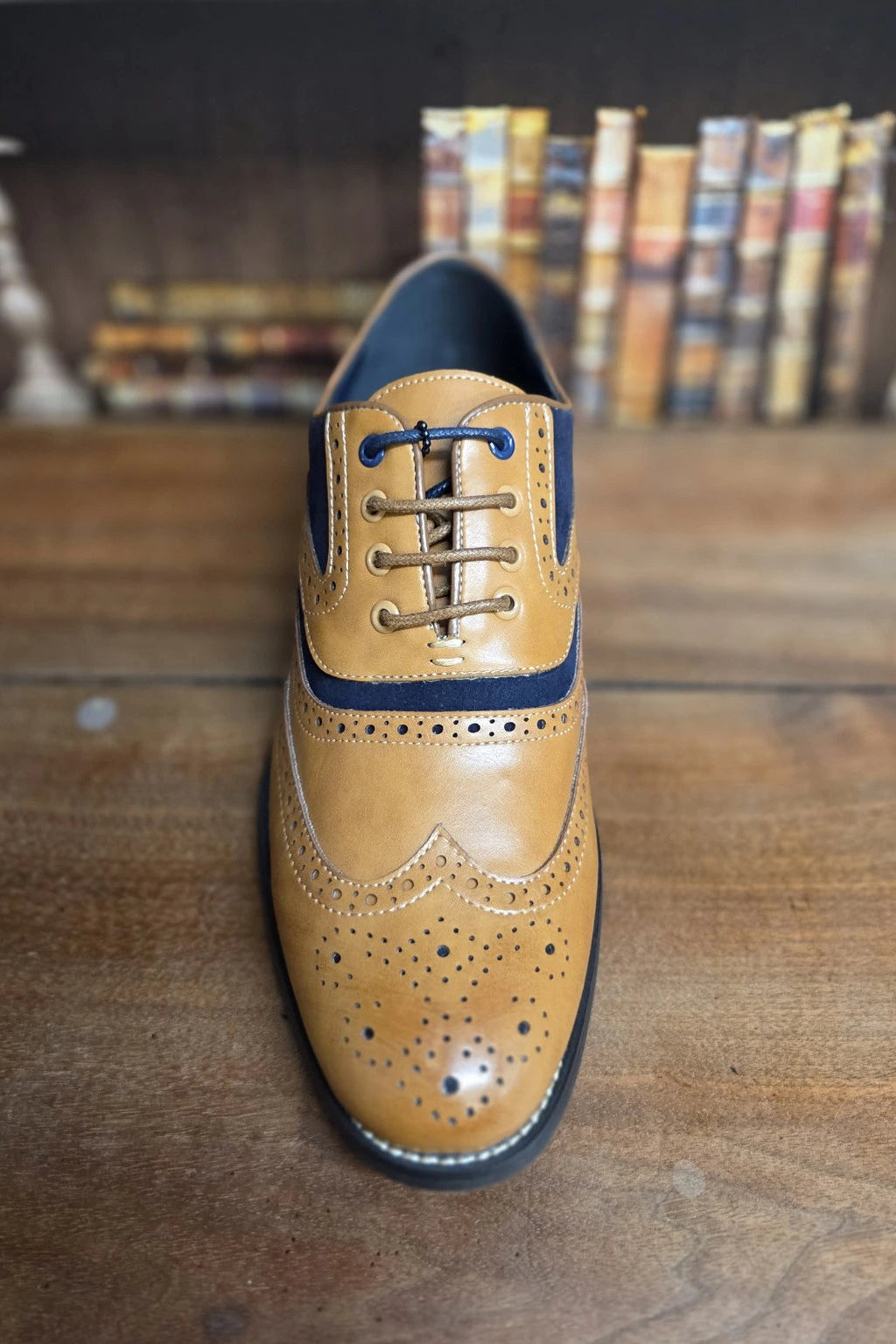 A tan and navy blue brogue wingtip dress shoe with laces, displayed on a wooden surface with a blurred background of books.