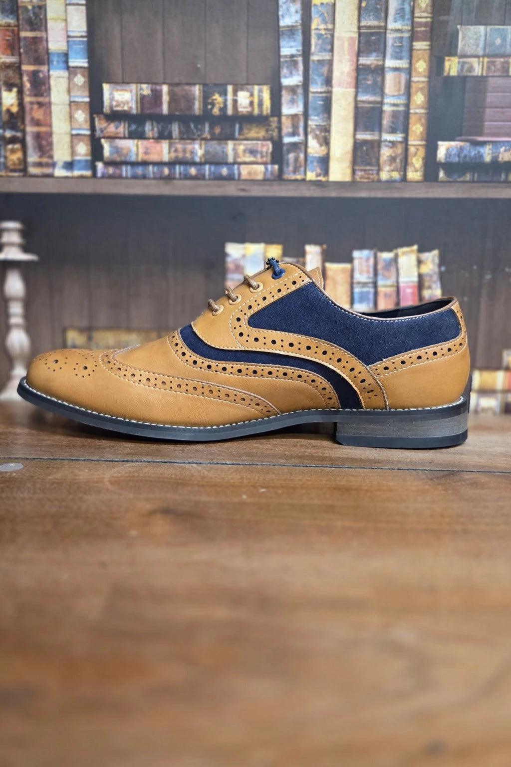 A men's wingtip brogue shoe in tan leather with navy blue suede overlays, resting on a wooden table with a blurred bookshelf background.