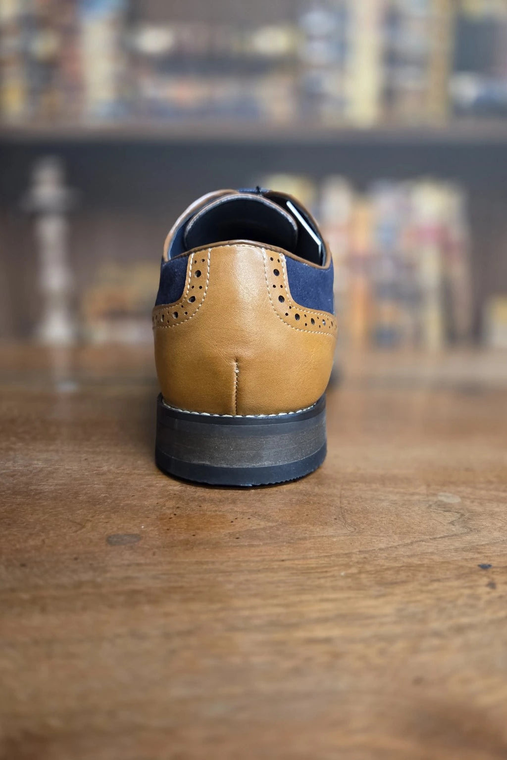A tan and navy blue leather wingtip shoe shown from the back, resting on a wooden table with a blurred bookshelf background.