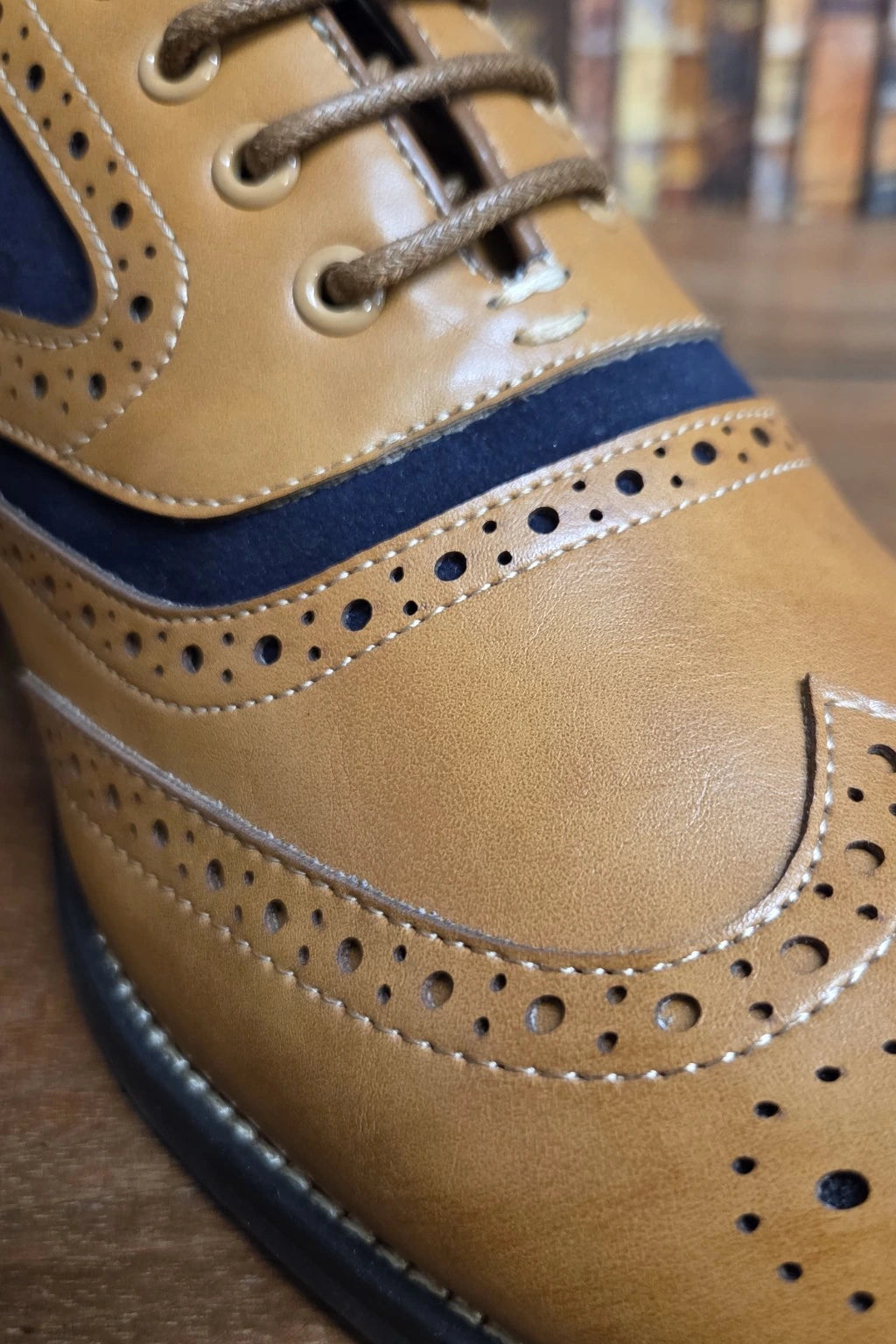 A close-up shot of a tan leather wingtip brogue shoe with dark navy suede panels and white contrast stitching, set against a blurred background of books on a wooden surface.
