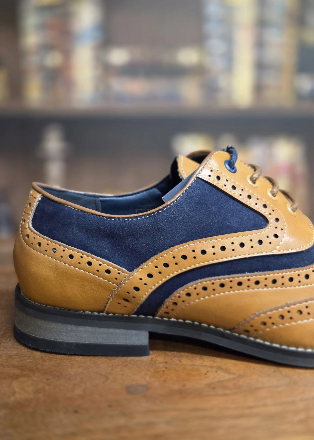 A tan and navy blue brogue wingtip shoe with perforated detailing, shown in a close-up side profile on a wooden surface with a blurred background.