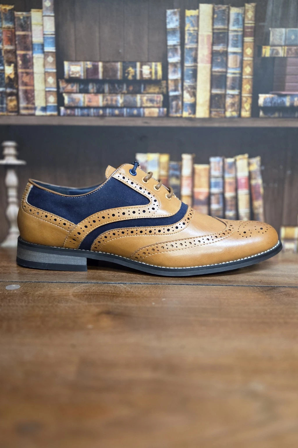 A men's wingtip oxford shoe in tan and navy blue leather, positioned on a wooden table with a blurred background of bookshelves.