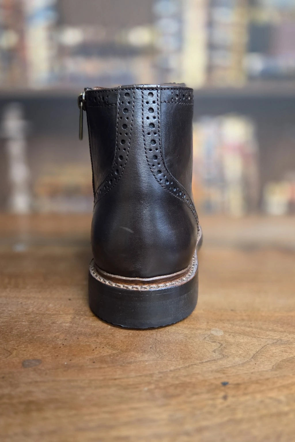 A single black leather ankle boot with brogue perforations and a side zipper, photographed standing upright on a wooden surface with a blurred indoor background.