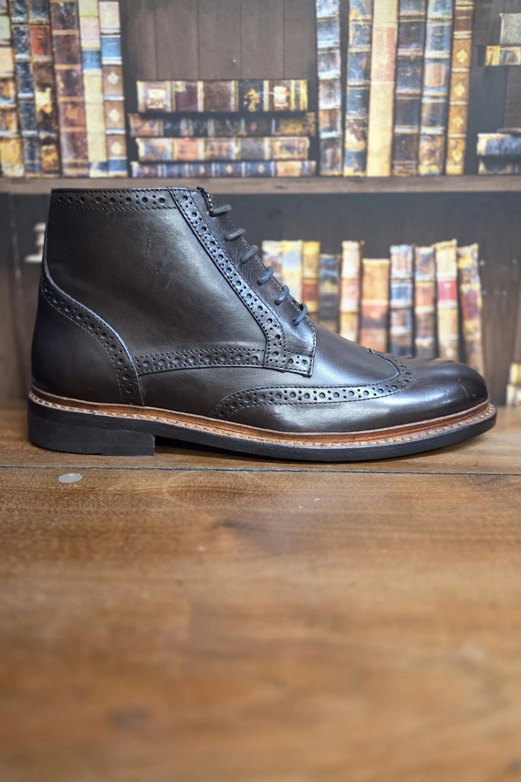A dark brown leather lace-up ankle boot with brogue detailing, positioned on a wooden table with a blurred bookshelf background.
