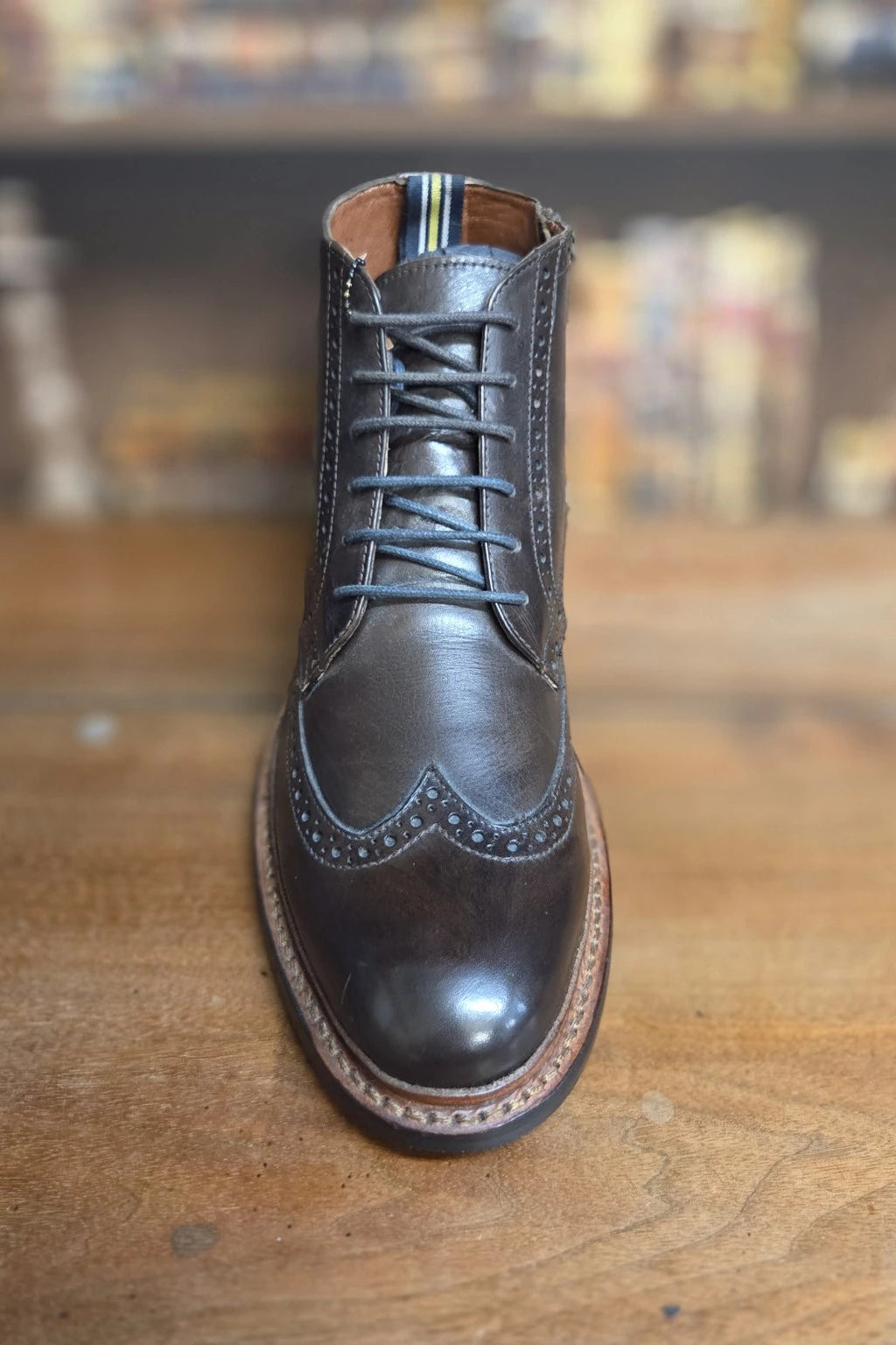 A dark brown leather lace-up wingtip boot with decorative brogue detailing, photographed from the front on a wooden surface with a blurred indoor background.