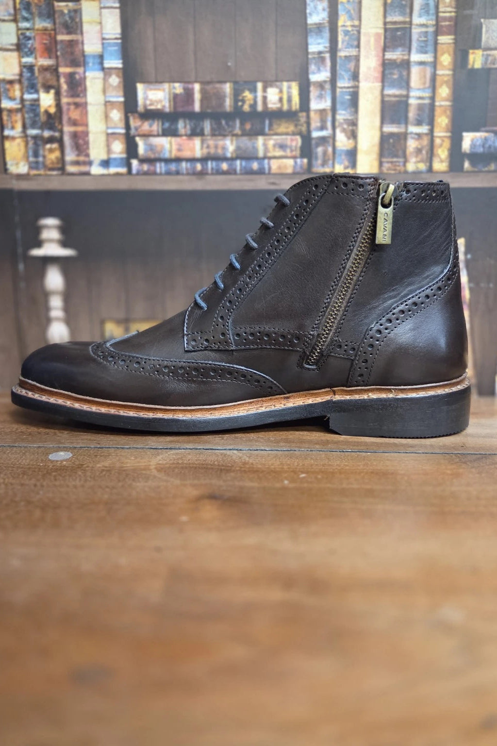A dark brown leather lace-up ankle boot with a side zipper, shown in profile on a wooden surface with a blurred bookshelf background.