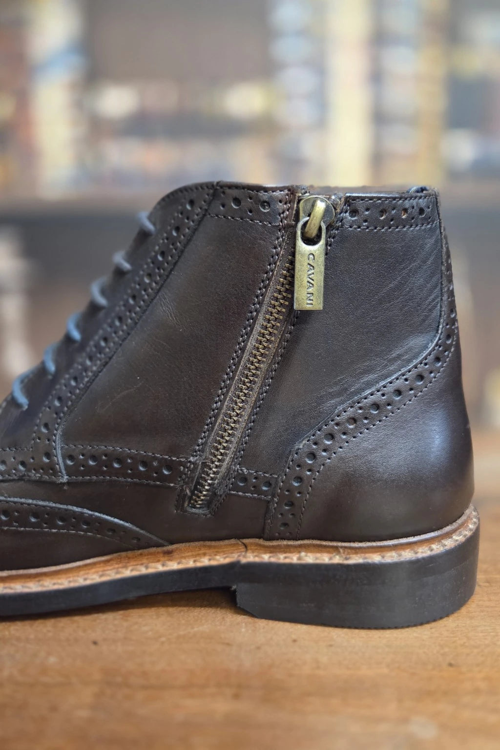 A close-up side view of a dark brown leather lace-up ankle boot with a side zipper, featuring a brass pull tab with the brand name 'Cavalli' and a tan midsole, resting on a wooden surface.