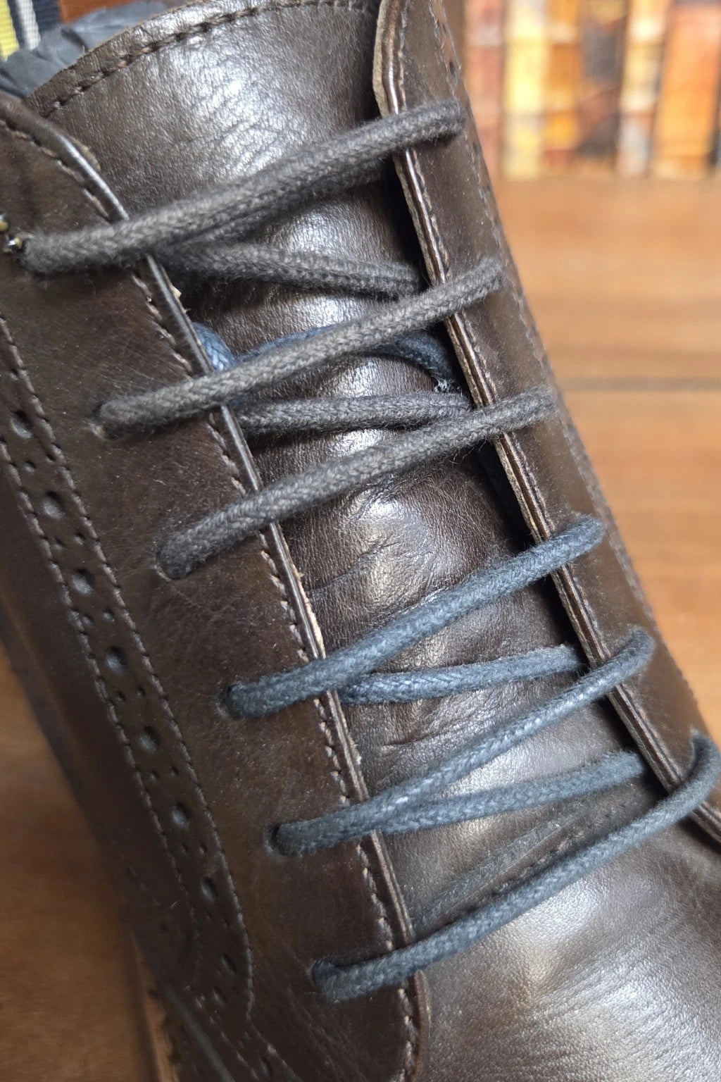 A close-up of a dark brown leather lace-up boot with dark gray laces, set against a blurred background of a wooden floor and bookshelves.