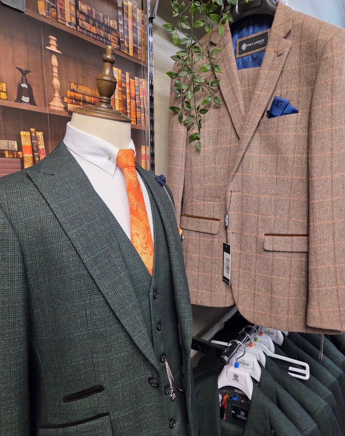 Two suits on mannequins with a Paul Andrew Harris brown checkered suit & a Cavani Caridi Olive Suit with an orange tie, against a bookshelf background.