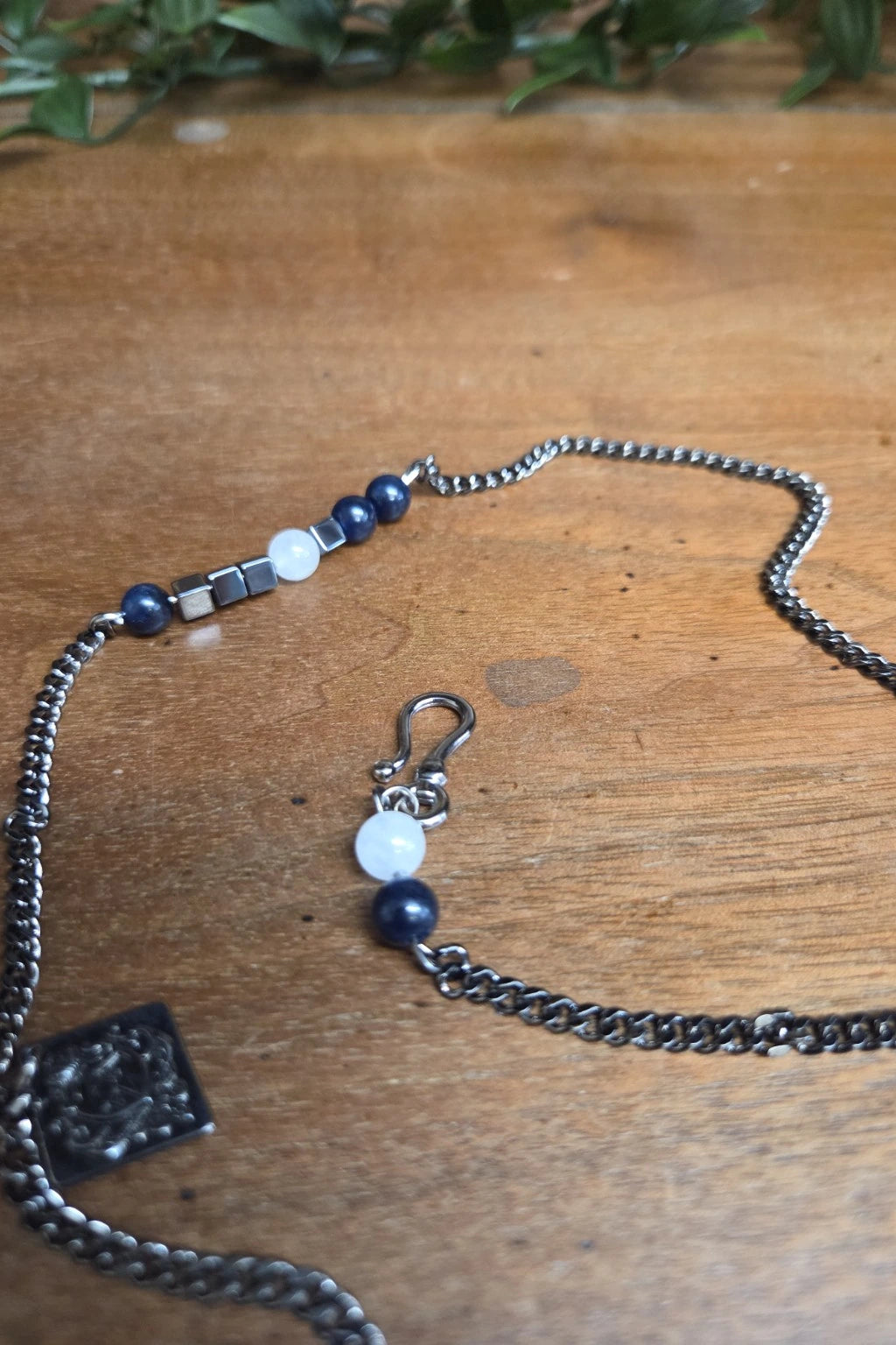 A close-up of the waistcoat chain laid on a wooden surface, featuring dark blue beads, a white round bead, silver-toned metal spacer, and a small dangling charm with a white bead and dark blue