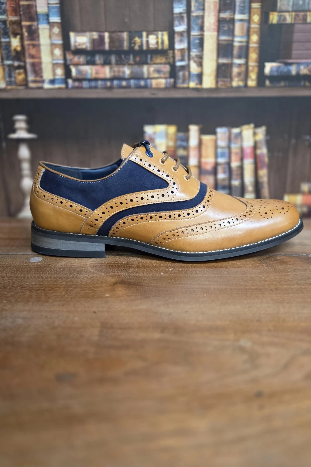 A men's wingtip brogue shoe in tan and navy blue, displayed on a wooden table with a blurred bookshelf background.