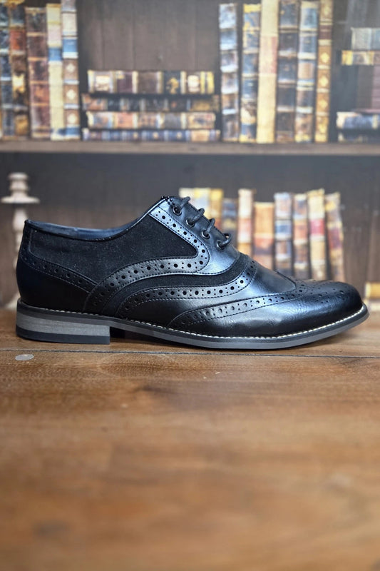 A single black leather wingtip brogue shoe with a suede overlay and lace-up closure, displayed on a wooden surface with a blurred bookshelf background.