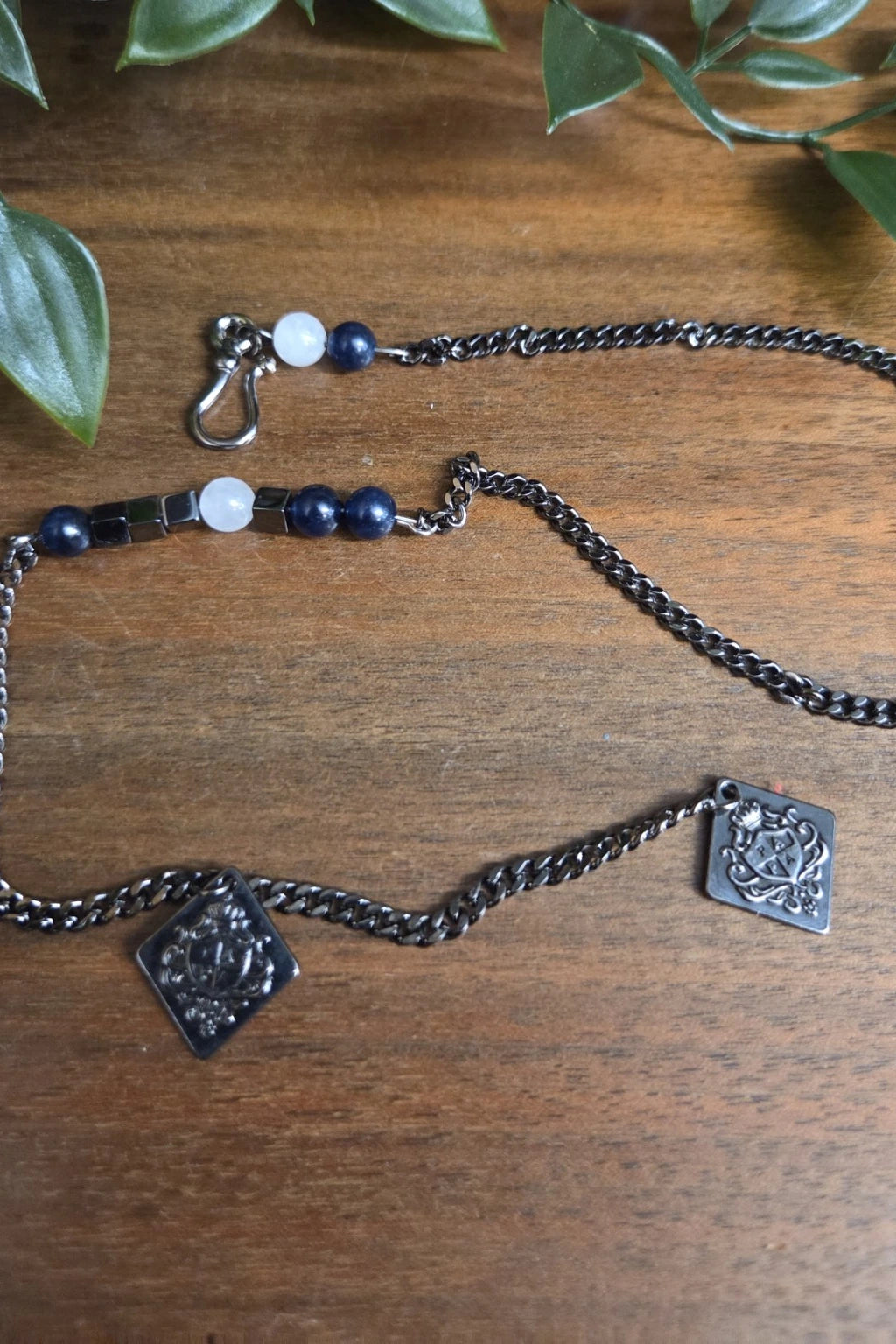 A dark gunmetal waistcoat chain with square engraved charms and white and navy beads, displayed on a wooden surface with green leaves in the background.